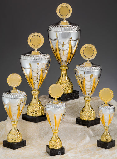 Eine Sammlung der **Pokale Bensheim 6-er Pokalserie 275 mm – 508 mm PK757910-6-E50** goldenen und silbernen Trophäen auf schwarzen Sockeln, unterschiedlicher Größe, arrangiert auf einem cremefarbenen Stoff.